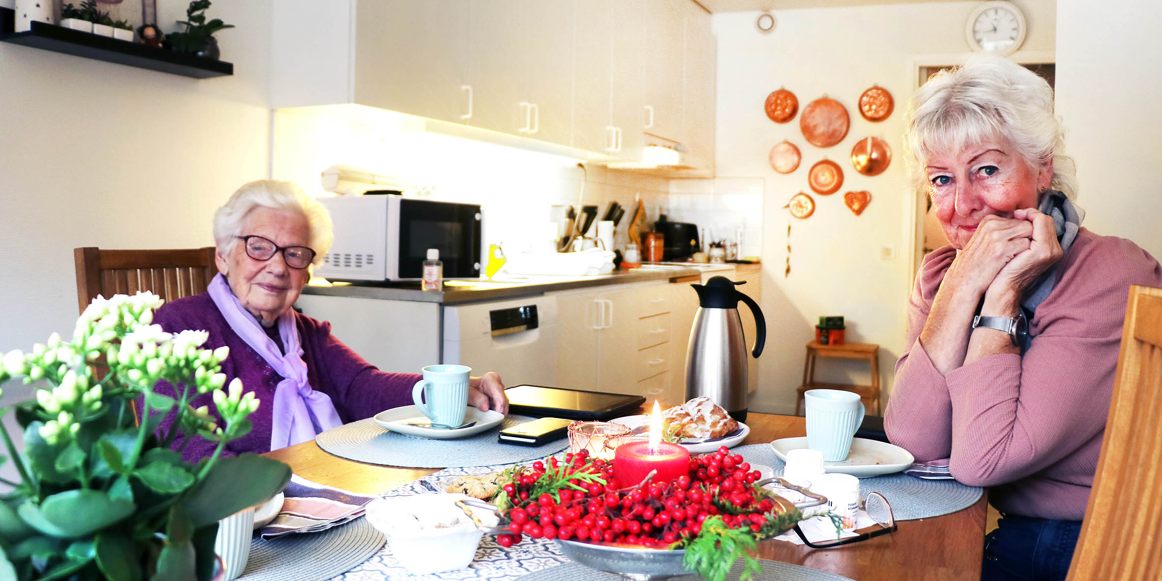 Två äldre kvinnor sitter vid sitt köksbord med kaffe och fikabröd på bordet och ler mot kameran.