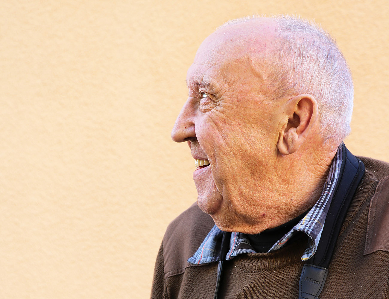 En äldre man med kort grått hår tittar åt vänster så man ser ansiktet i profil. Han har på sig en brun tröja och en blå skjorta undertill.