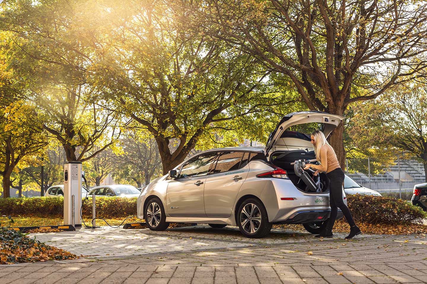 En gråmetallic bil står parkerad vid en laddningsstation. En ljushårig tjej lägger in en barnvagn i bagaget