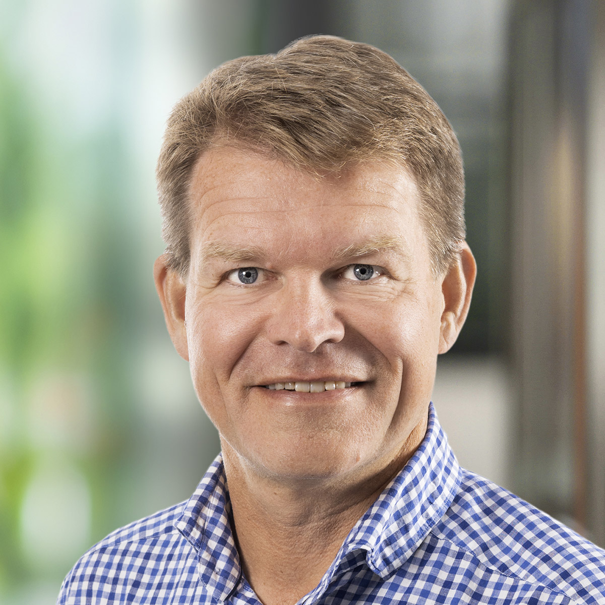 En man med kort ljust hår med lite sned bena och blå vit rutig skjorta tittar in i kameran och ler