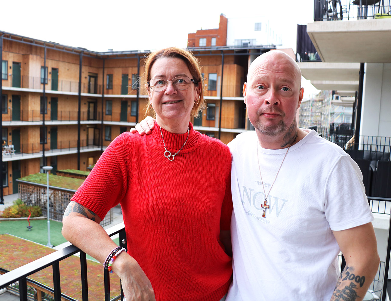 En kvinna med en röd polotröja och en man med en vit t-shirt står på en balkong och håller armen om varandra. I bakgrunden ser man en innergård och en fasad med balkonger.