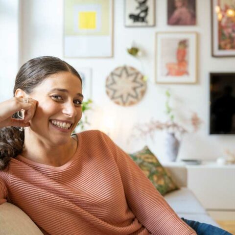 En kvinna med rosa tröja sitter i en soffa och tittar in i kameran och ler.