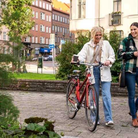 Två kvinnor promenerar i stadsmiljö, en av dem leder en cykel