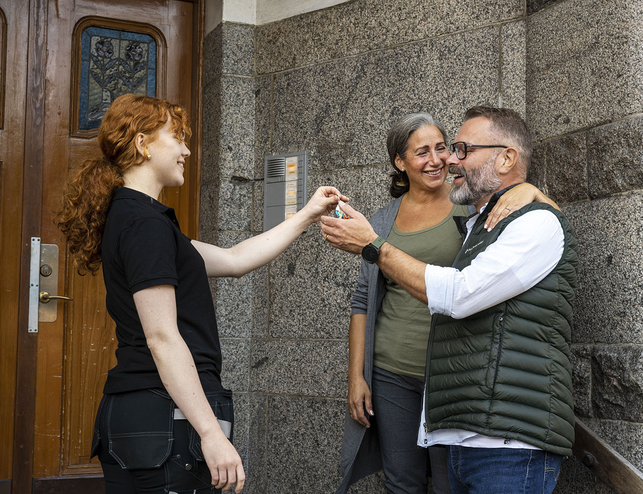 En rödhårig kvinna överlämnar en nyckel till ett par som står utanför en dörrport.