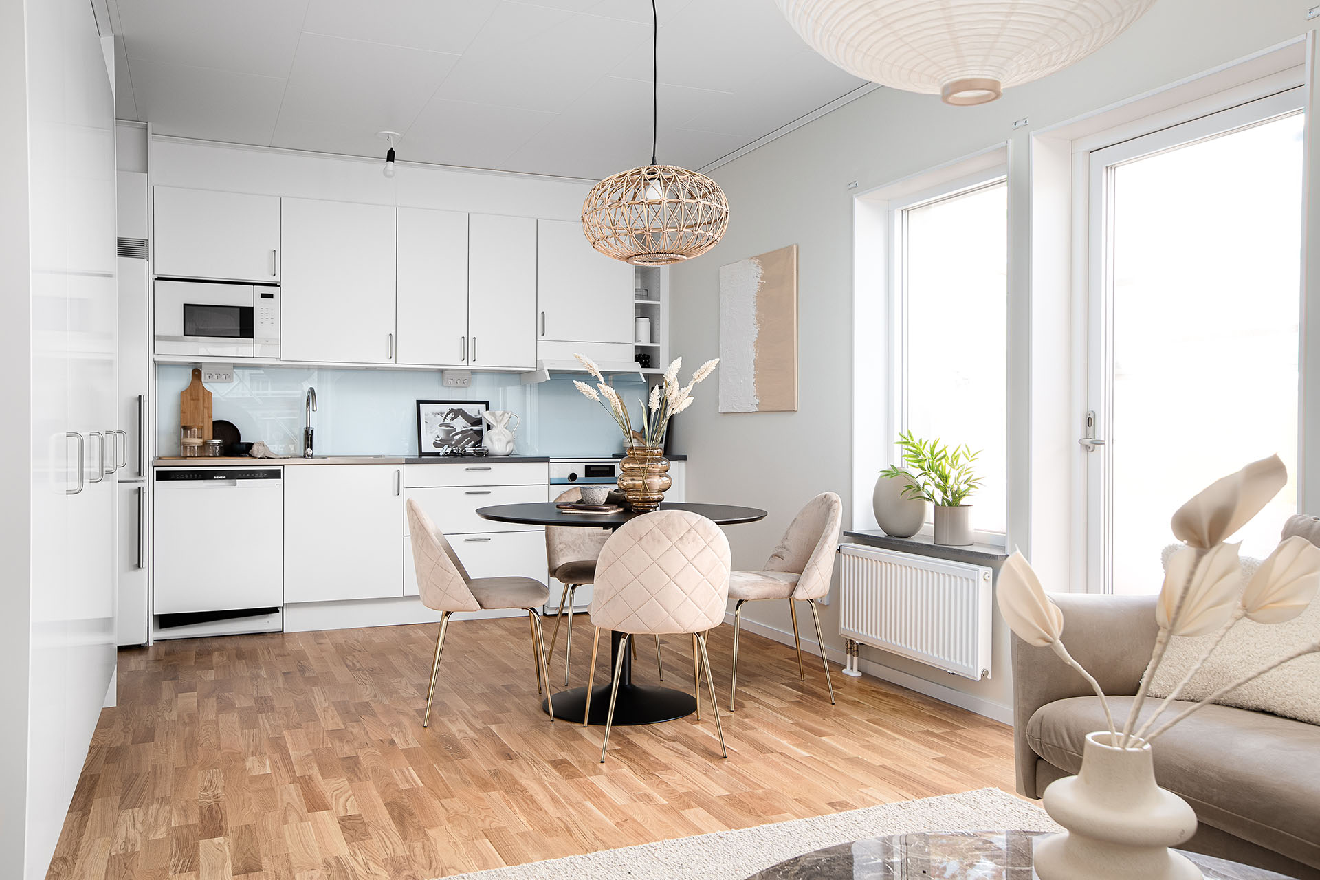 Ett vitt kök och kombinerat vardagsrum. Köket har vita skåpluckor och på det ljusa trägolvet står det ett runt köksbord med tygstolar runt om.