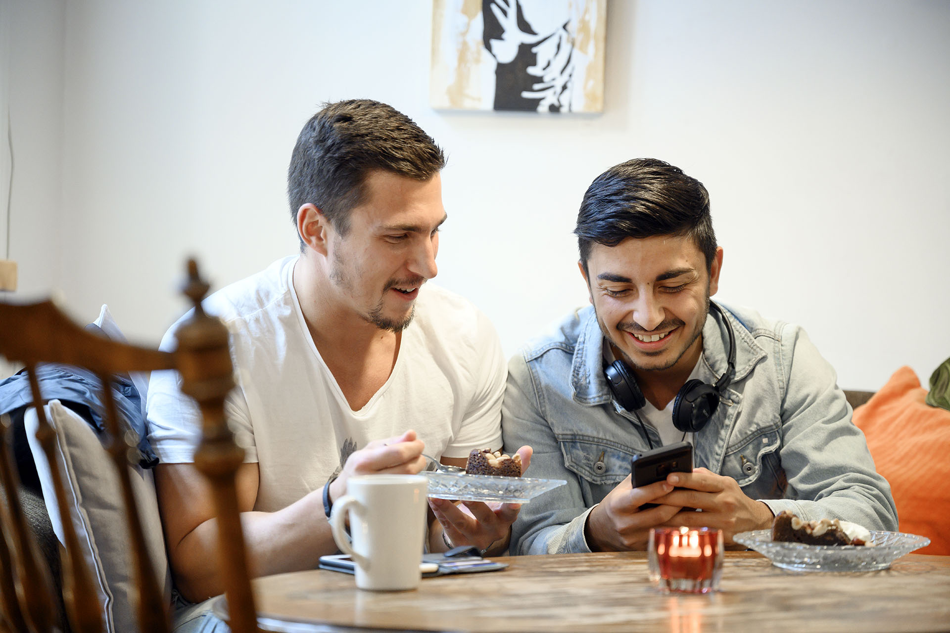 Två killar, den ena klädd i en vit tshirt och den andre i en jeansjacka sitter på ett cafe och dricker kaffe och tittar på sin mobiltelefon