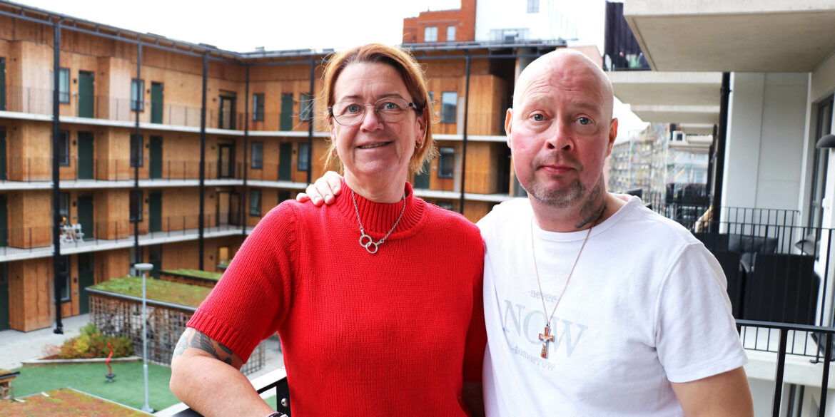 En kvinna med en röd polotröja och en man med en vit t-shirt står på en balkong och håller armen om varandra. I bakgrunden ser man en innergård och en fasad med balkonger.