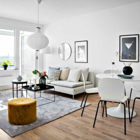 Ett vardagsrum med en beige soffa, en stor fyrkantig matta och en gul sittpuff på golvet