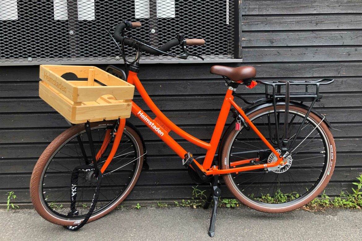En orange elcykel som har en stor träkorg framtill står parkerad mot en svart vägg