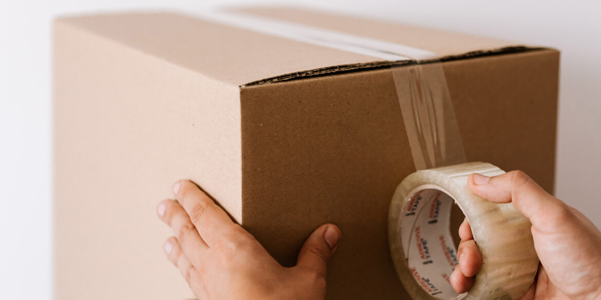 En närbild på en stor brun kartong och där man ser två händer som drar en bred tejp över lådan.