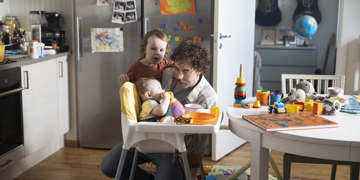 En pappa med mörkt kort hår står på huk framför sina två barn som sitter vid köksbordet. På bordet ser man leksaker och frukostsaker som står framme.