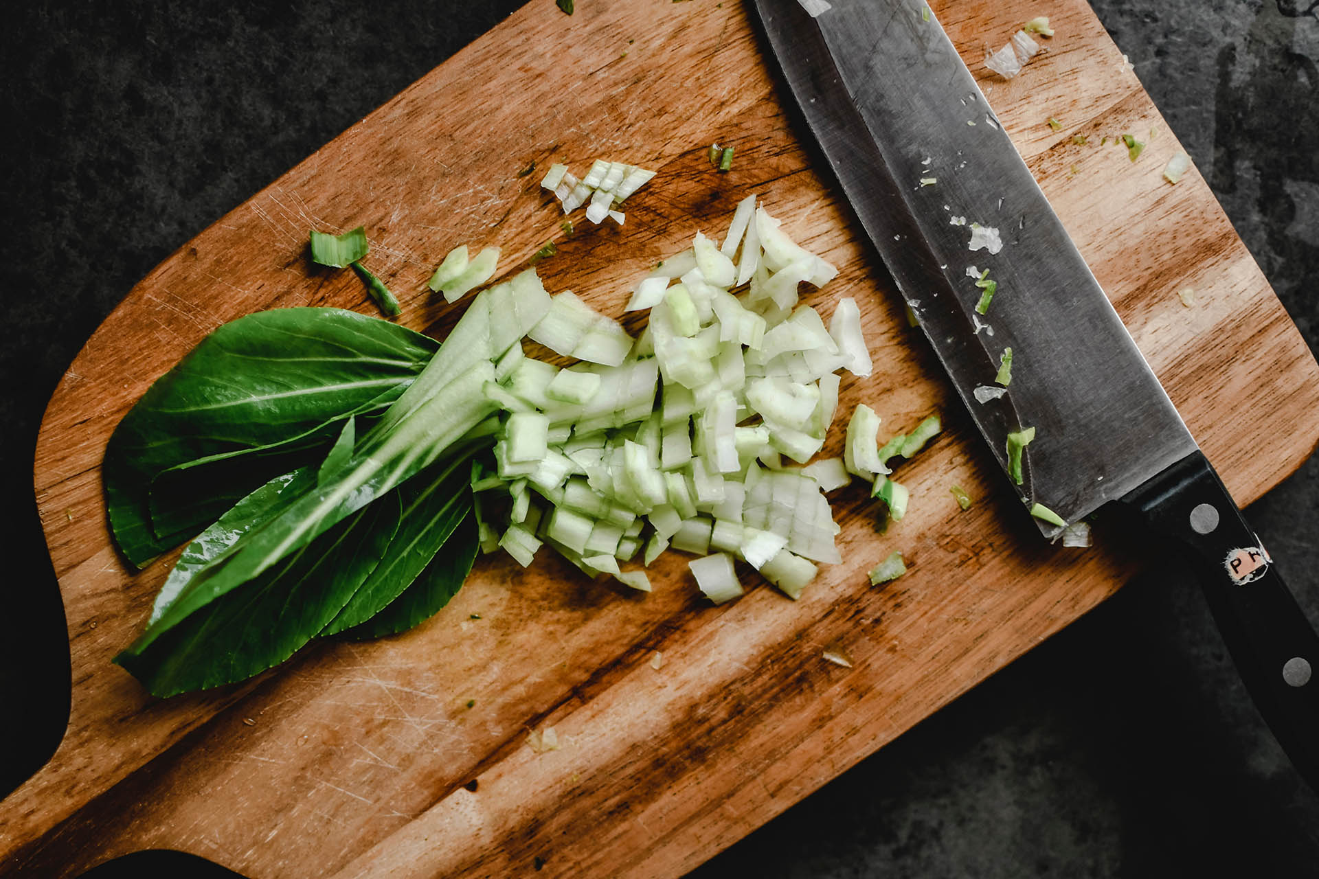 En skärbräda i trä som det ligger en stor kniv på och en grön grönsak som är till hälften finhackad.