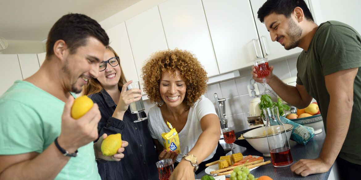 Två tjejer och två killar står i ett kök och skrattar. I händerna håller de citroner, kex och glas med dryck i.