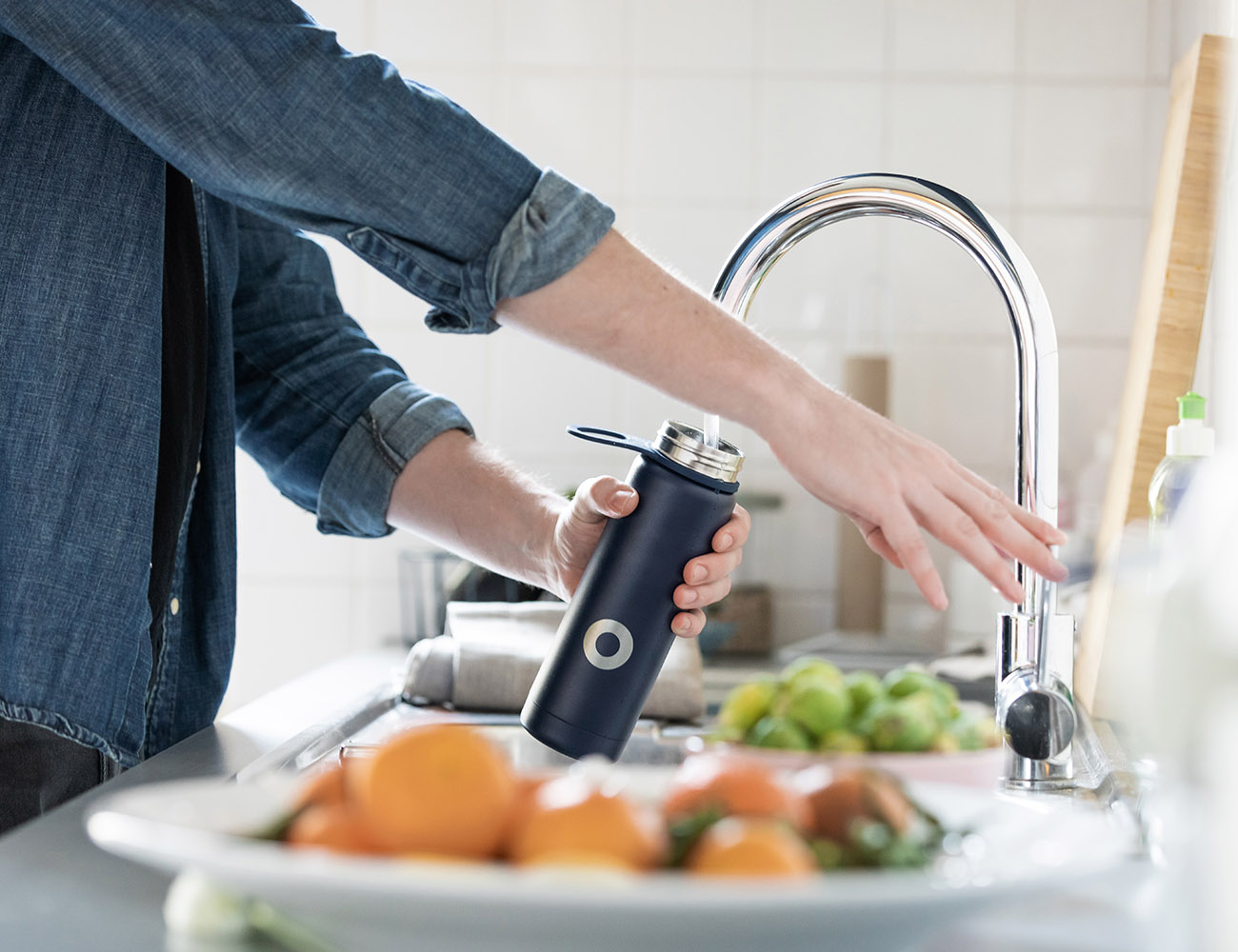 En person häller upp vatten i en svart flaska. Framför ser man ett fat med apelsiner