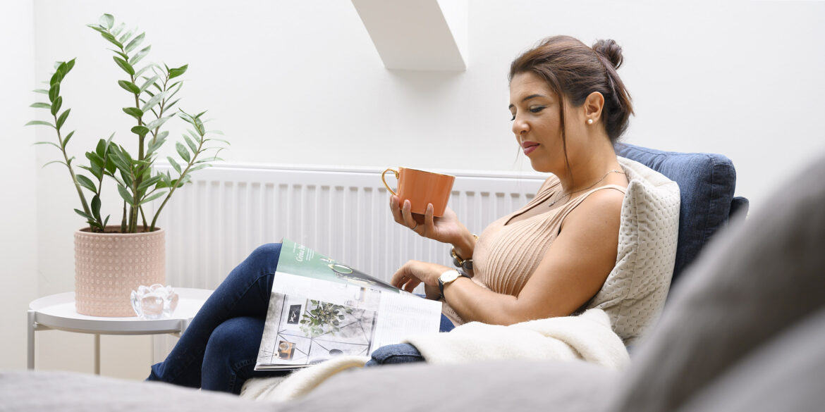 En kvinna sitter i en ljus soffa med en orange kaffekopp i handen och en tidning i knät som hon läser