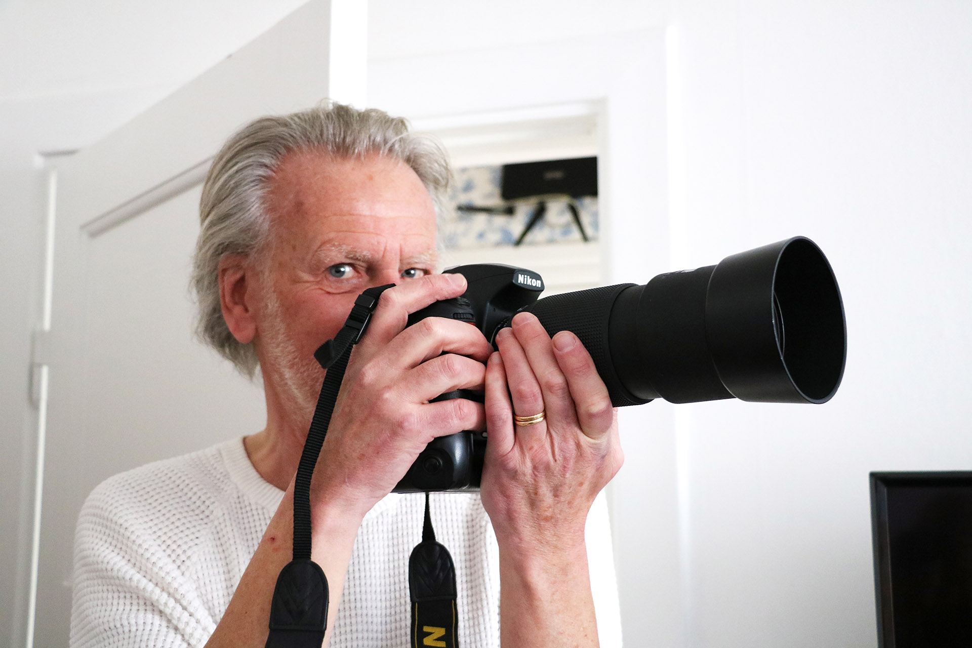 En man med grått och hår och en vit farfarströja håller en kamera i handen och kikar