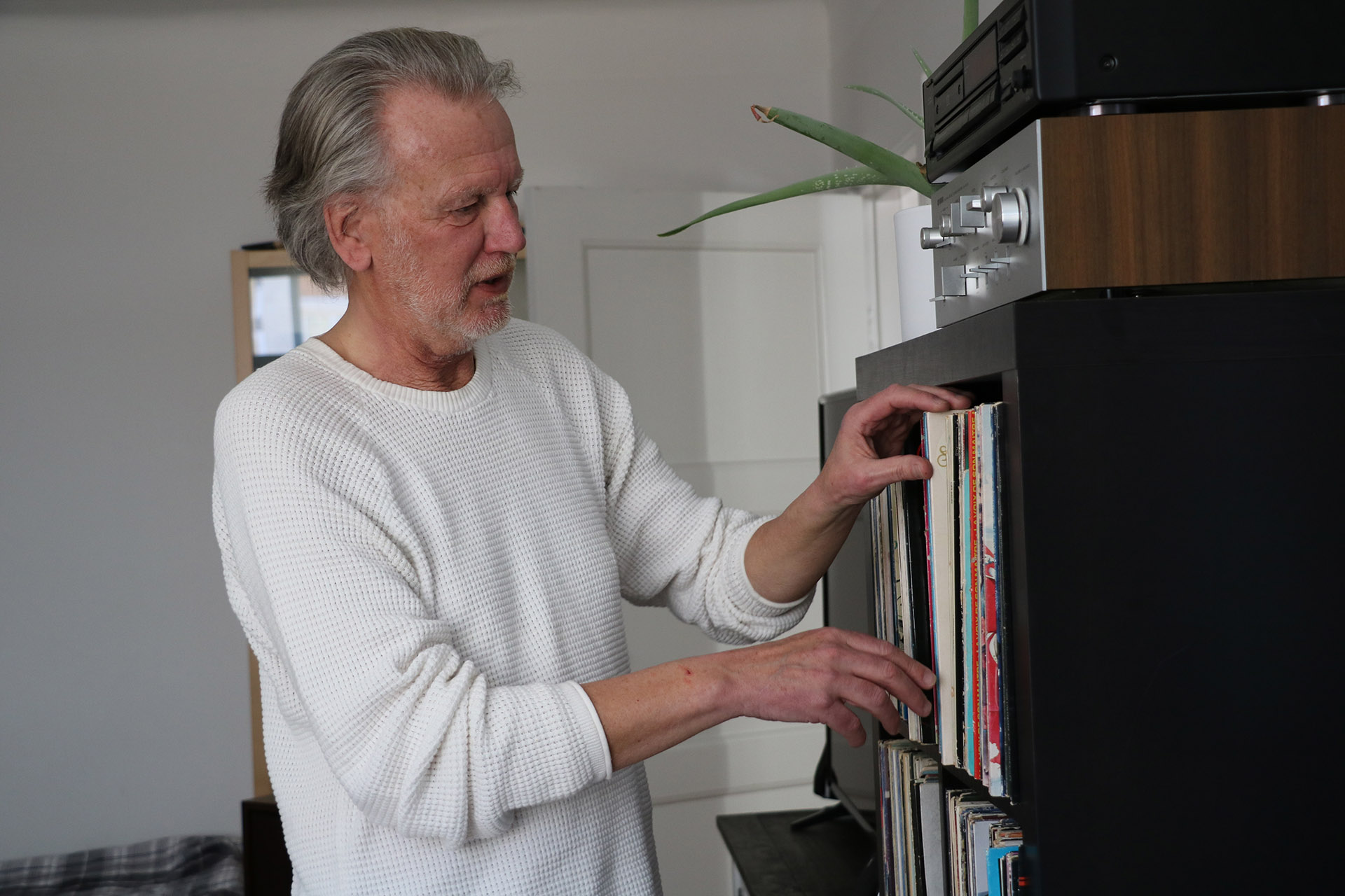 En man med grått hår och vit tröja står framför sin samling av LP skivor och kollar