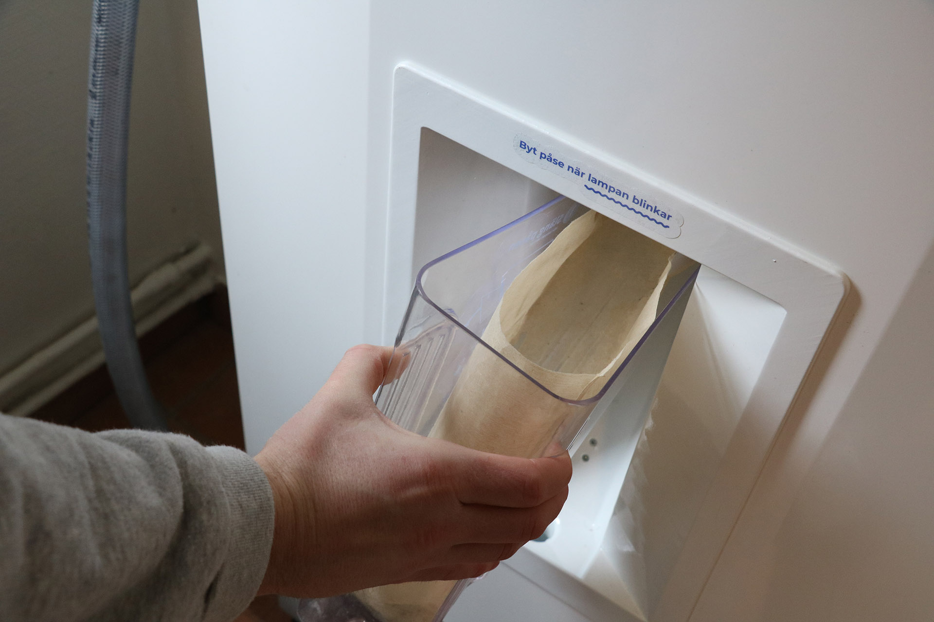 En närbild på en hand som håller i en behållare mot en vit maskin. På maskinen står texten; byt påse när lampan blinkar