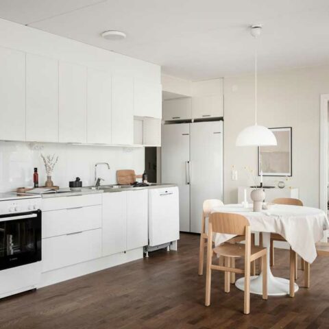 Ett vitt kök med ett mörkt trägolv. Står ett litet runt matbord med tre trästolar framför