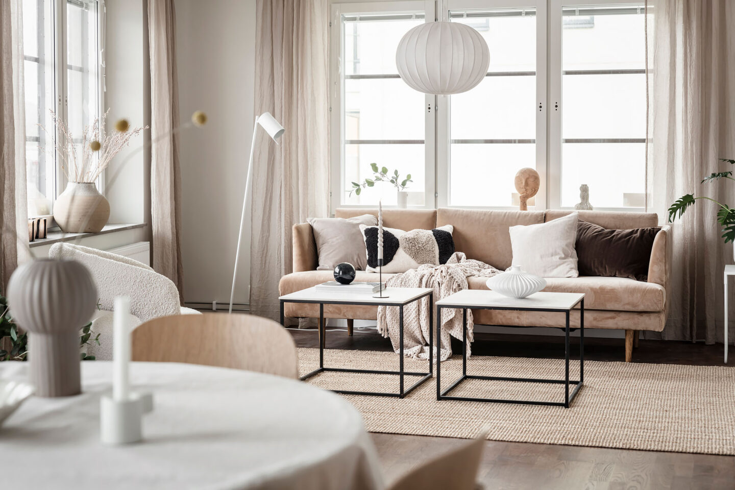 I förgrunden ser man ett köksbord, bortom köksbordet ser man ett vardagsrum med soffa, bord och fönster i bakgrunden