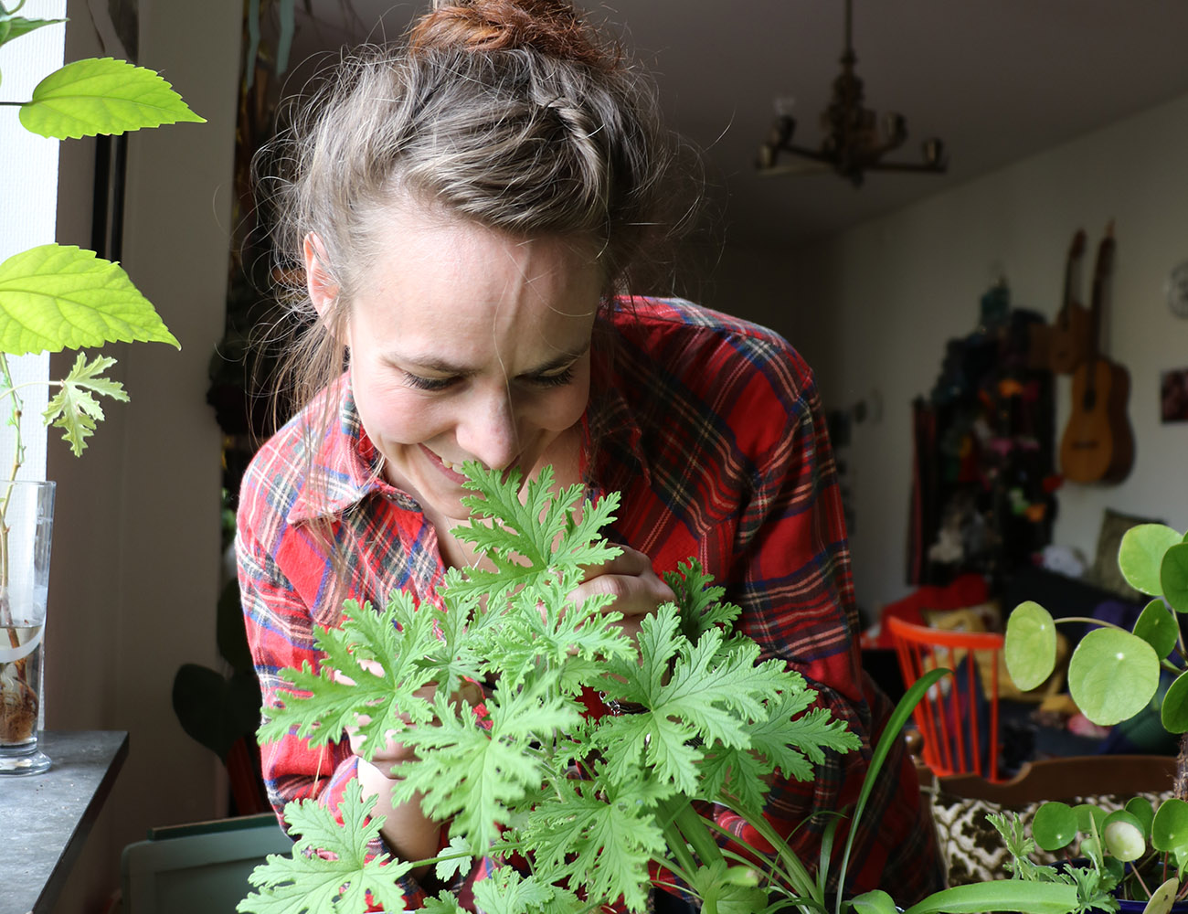 En kvinna med mörkt hår och rödrutig skjorta sitter vid sitt köksbord och tittar på sina gröna växter i fönsterkarmen.