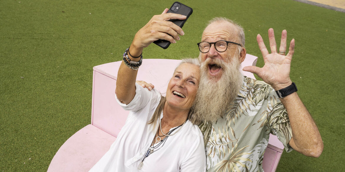 En äldre man med vitt stort skägg och en kvinna med en vit tunika sitter på en rosa bänk och tar en selfie och skrattar