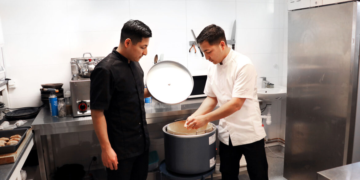 Två killar i kocktröjor står vid en stor riskastrull i ett kök