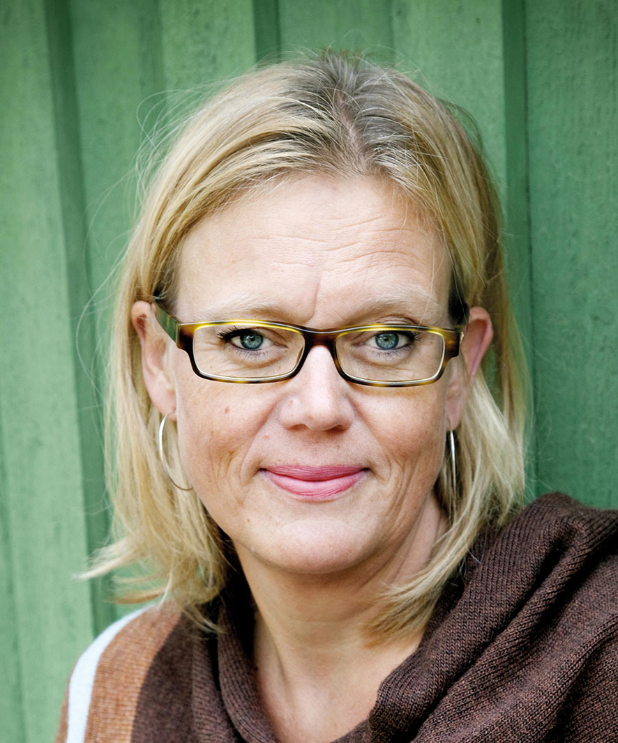 En blond kvinna med en brun tröja och glasögon tittar rakt in i kameran