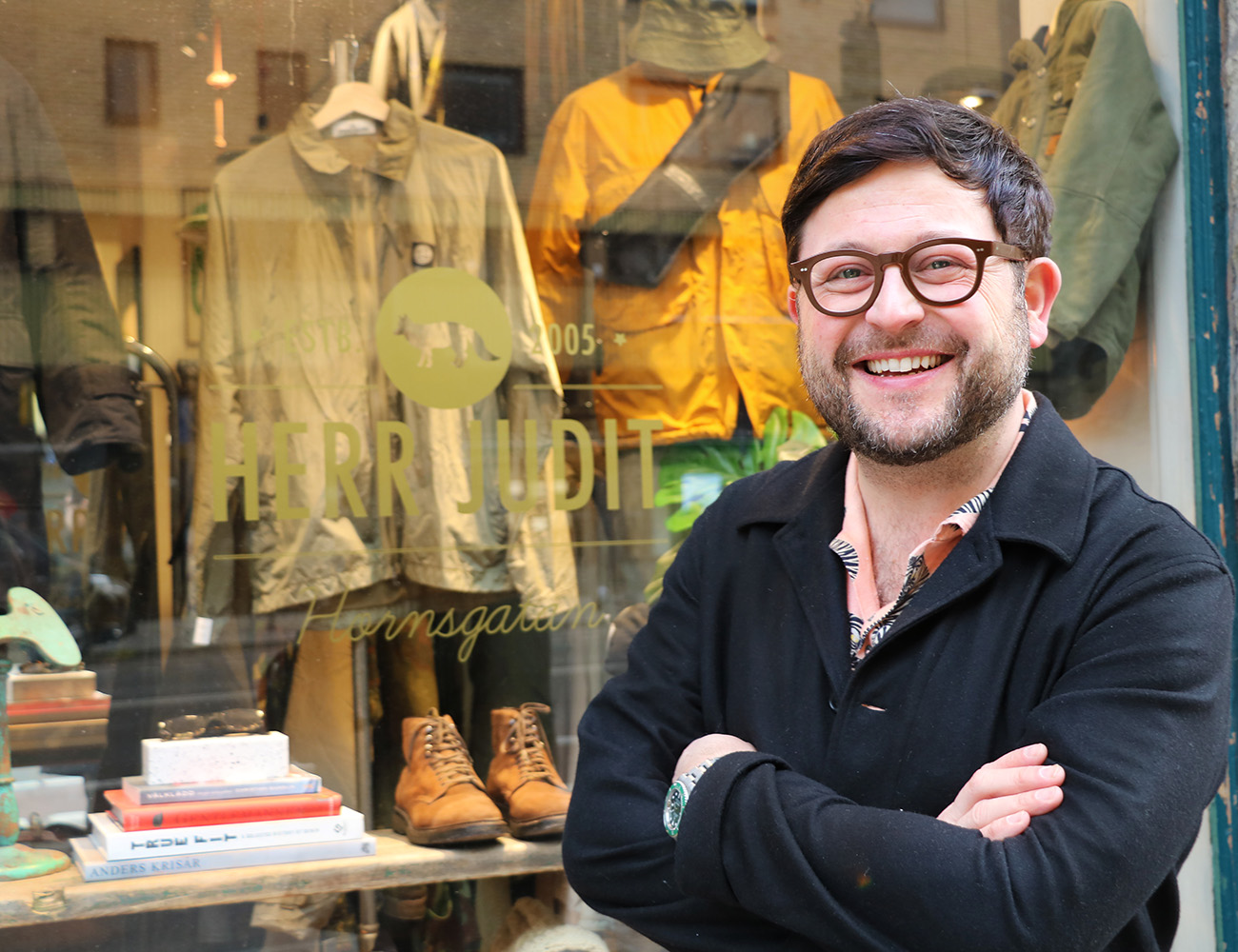 En man med glasögon och en svart skjorta står framför ett skyltfönster som det hänger kläder i