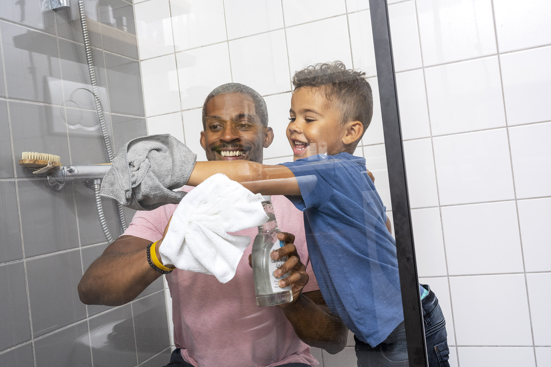 En pappa och son står i ett duschutrymme och städar väggen med varsin trasa