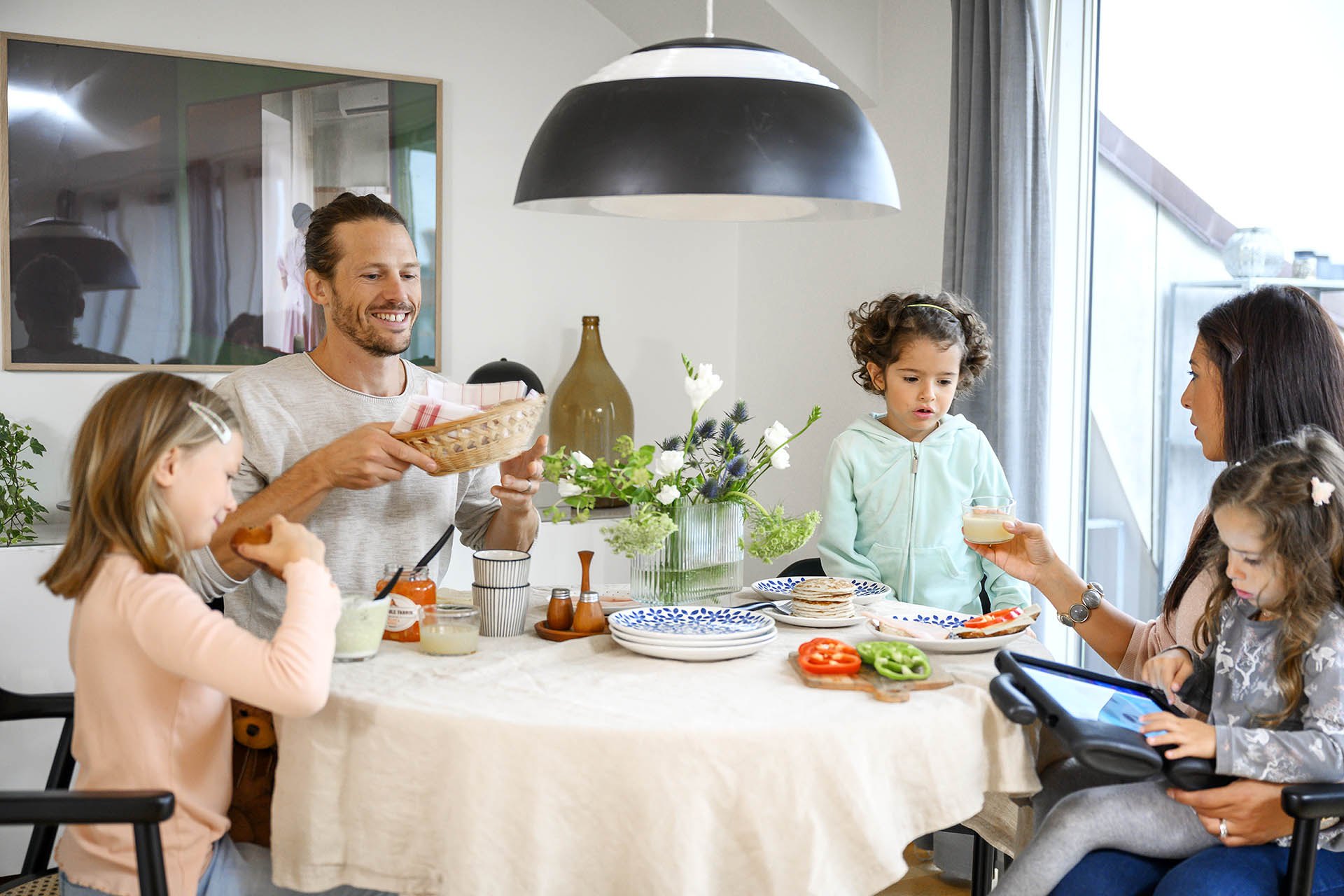 En familj med två barn sitter runt ett köksbord där det står porslin, mat och en stor blomvas på bordet