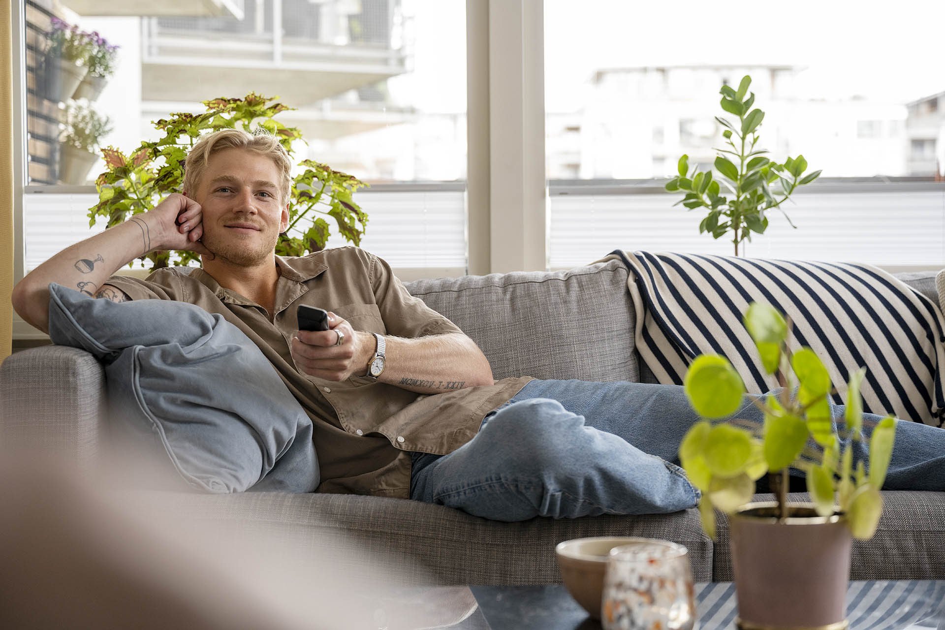 En kille ligger i en grå soffa med massa kuddar i och håller i en fjärrkontroll som han pekar mot kamera