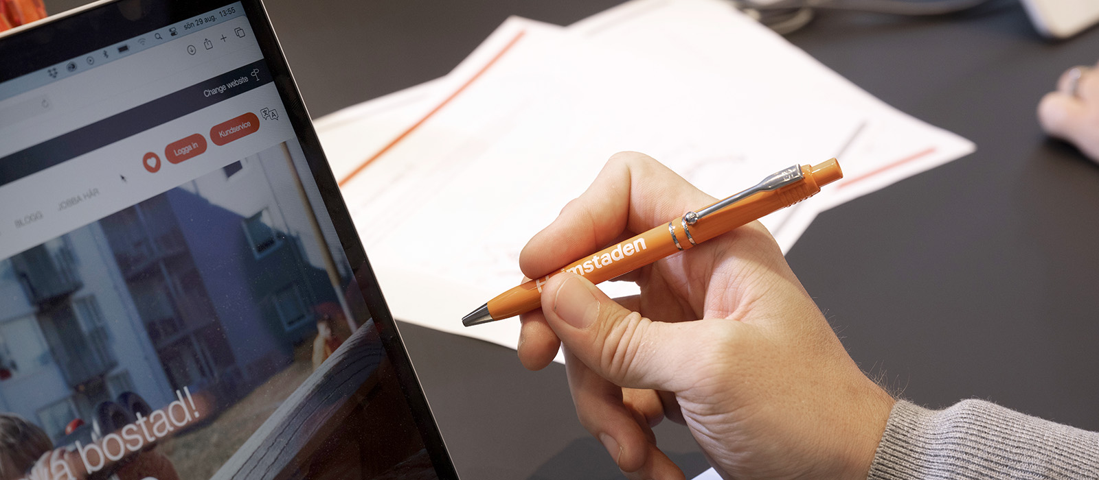En närbild på en hand som håller i en orange penna. En dator och vita papper ligger på skrivbordet.