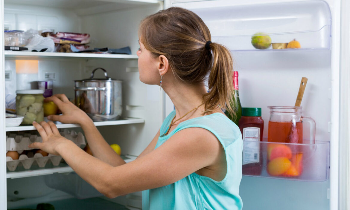 En kvinna med turkos tröja står och tittar in i sitt kylskåp