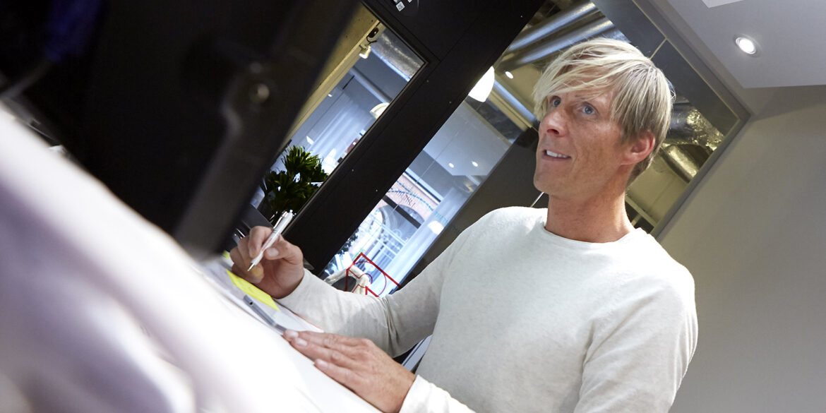 En kille i vit tröja står vid ett skrivbord och skriver på ett papper.