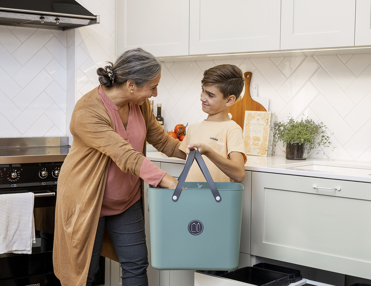 En kvinna med en lång beige kofta och en pojke med en gul tröja, står i ett kök och håller i en turkos plastlåda med handtag.