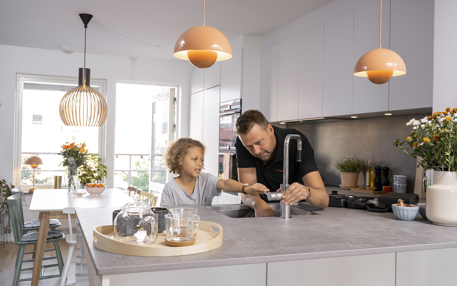 En man med arbetskläder och en pojke står vid en kökskran i köket