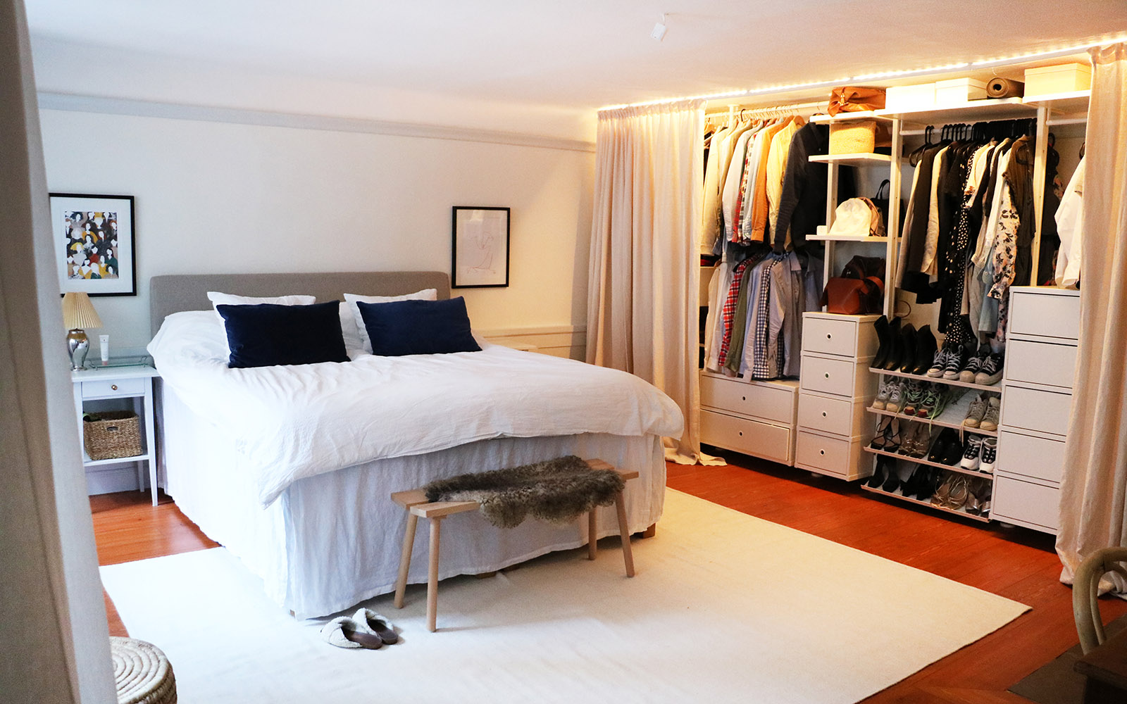 Ett sovrum med en stor säng med vitt överkast. En garderob längs hela väggen är öppen och man ser kläder och skor i den.