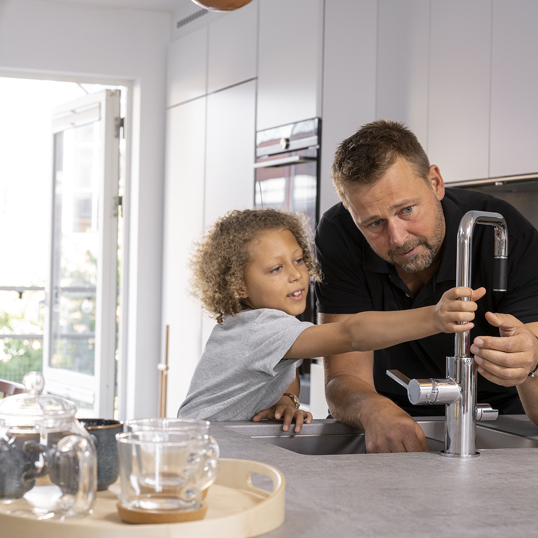 En man med svart pikétröja och ett barn med en grå t-shirt står vid ett vattenkran i köket.