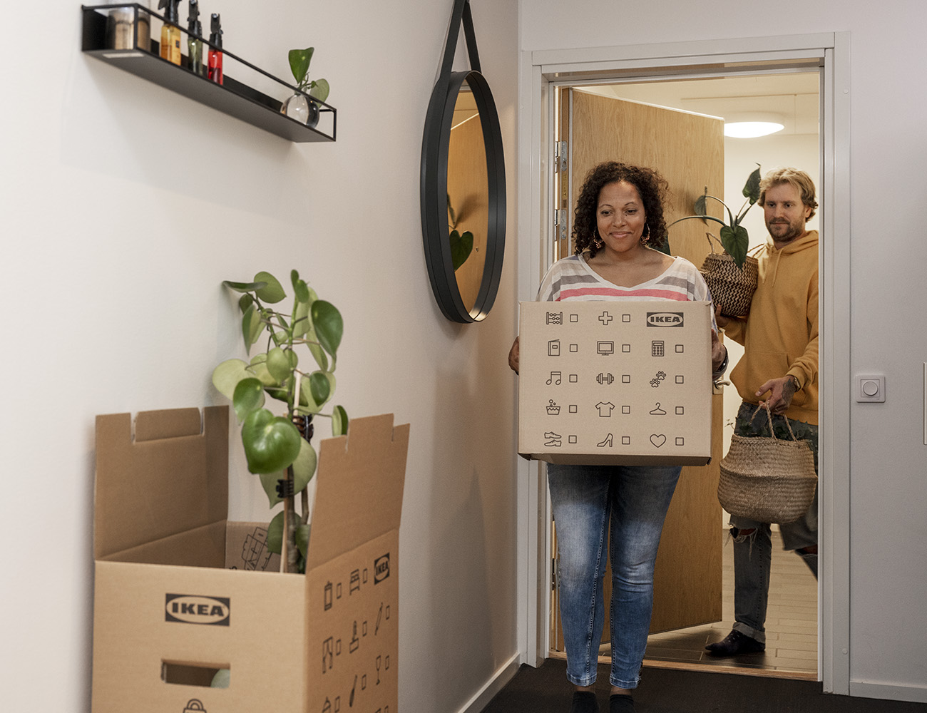 En kvinna kommer in i en bostad med en stor flyttkartong i famnen. En man kommer bakom bärandes på en grön växt.