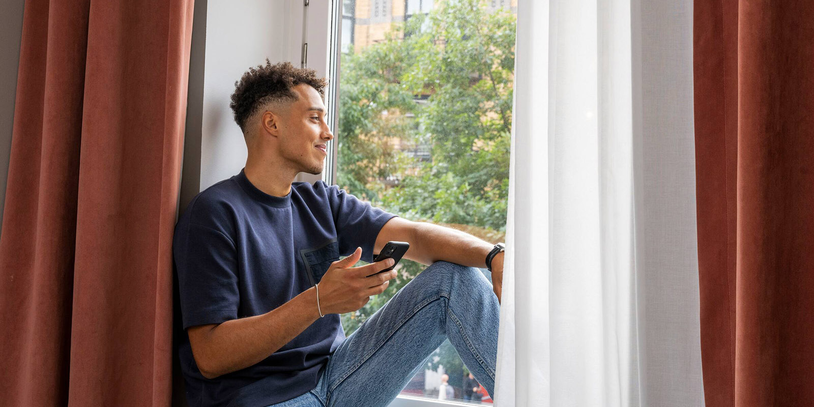 En ung kille med jeans och blå t-shirt, sitter i fönstret med sin mobil i handen och tittar ut.