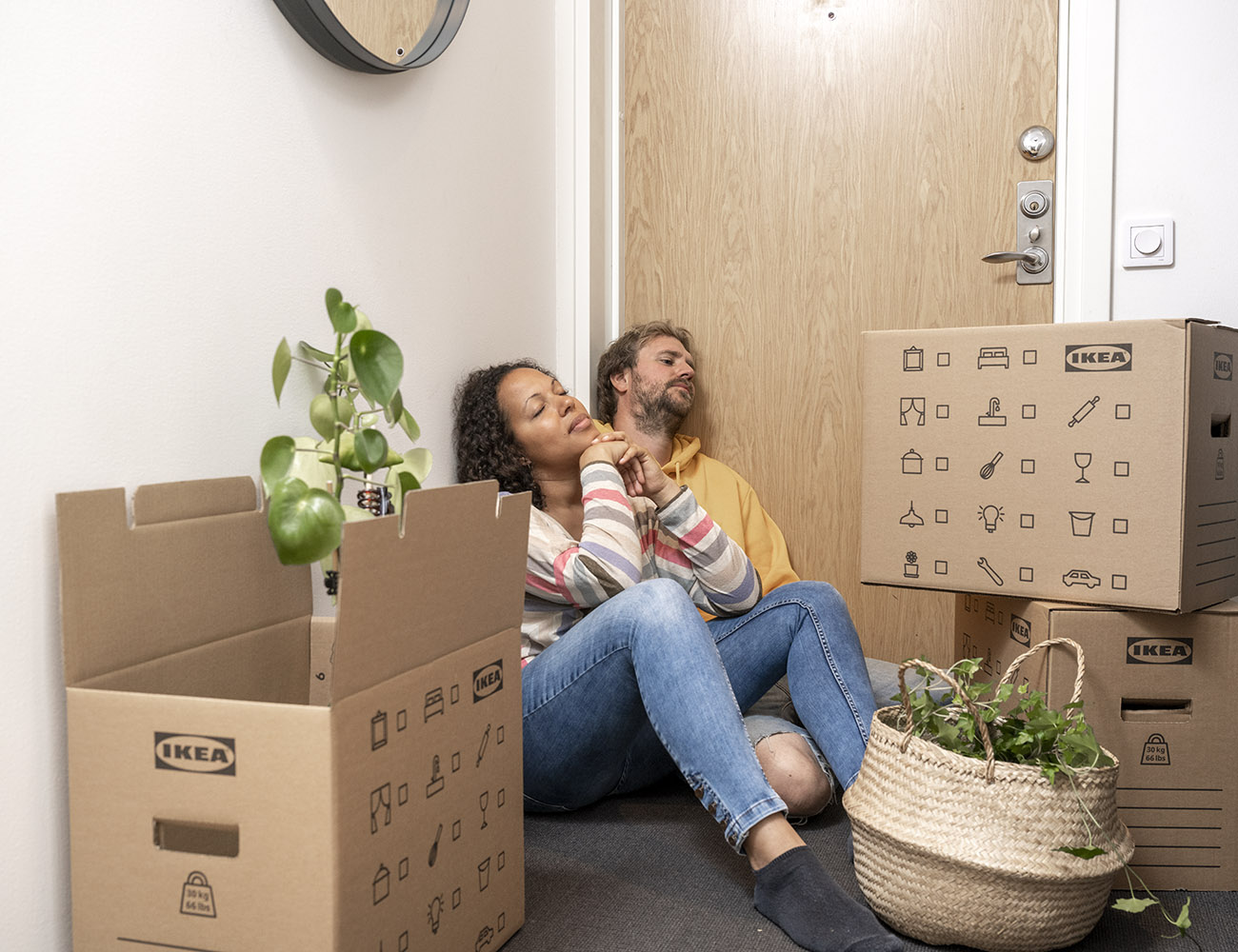 En kvinna och en man sitter på golvet framför sin ytterdörr. På golvet står det flyttkartonger med gröna växter i.