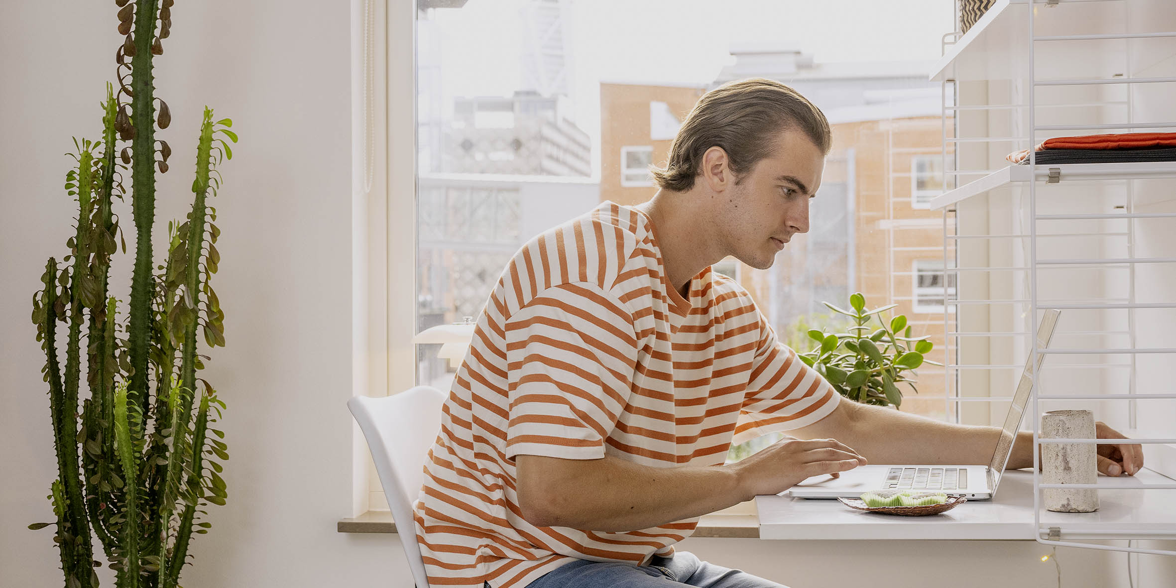 En ung kille med randig tshirt sitter vid sitt skrivbord och tittar på en laptop.