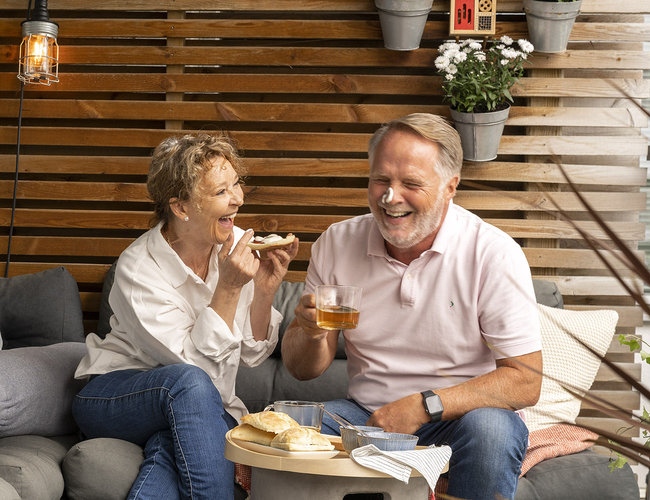 En man och kvinna som är somrigt klädda, sitter på en balkong och fikar smörgåsar och te. Mannen har fått en klick smör på sin näsa som de skrattar åt.