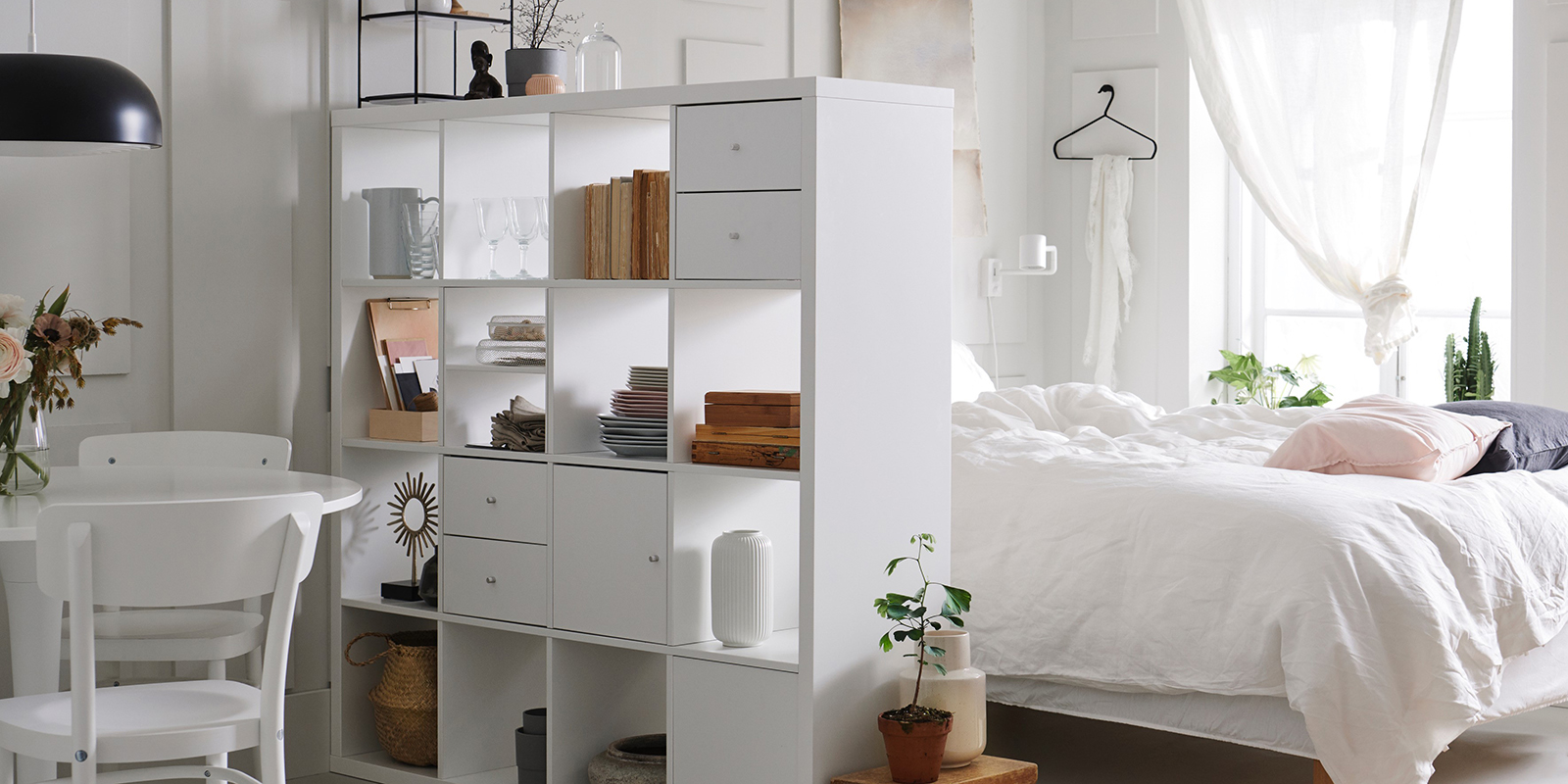 Ett rum med en säng bäddat i vita textilier och en vit bokhylla står.