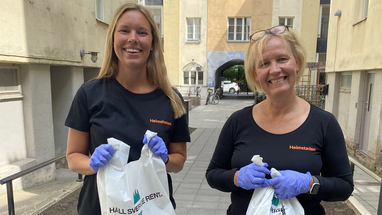 Två kvinnor med svarta tröjor som det står Heimstaden på, står med en påse i handen som det står Håll Sverige rent på.