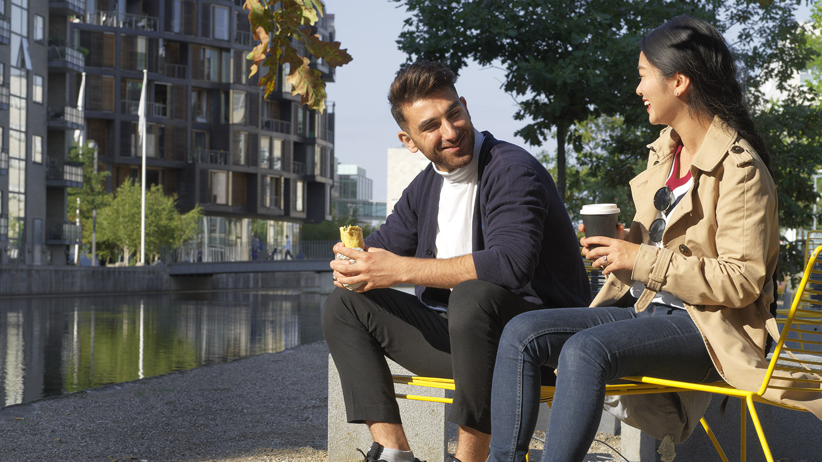 Ett ungt par sitter vid en kanal där man ser ett svart höghus bakom. De sitter på ett par gula stolar och dricker kaffe och pratar.