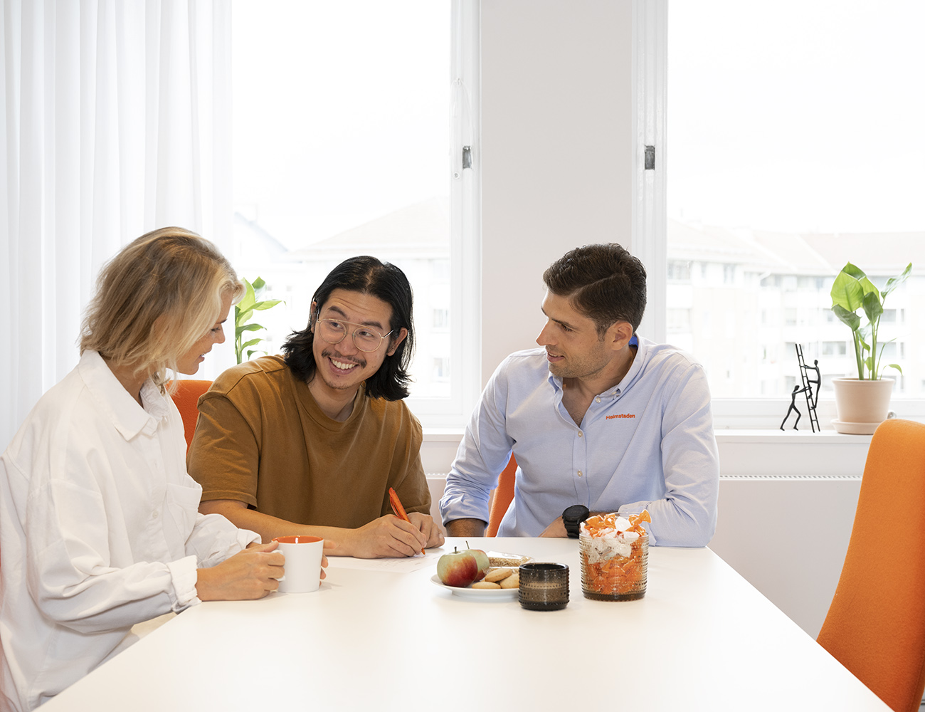 En kvinna i vit skjorta sitter tillsammans med två killar i blå skjorta och brun tröja vid ett konferensbord och skriver på ett papper.
