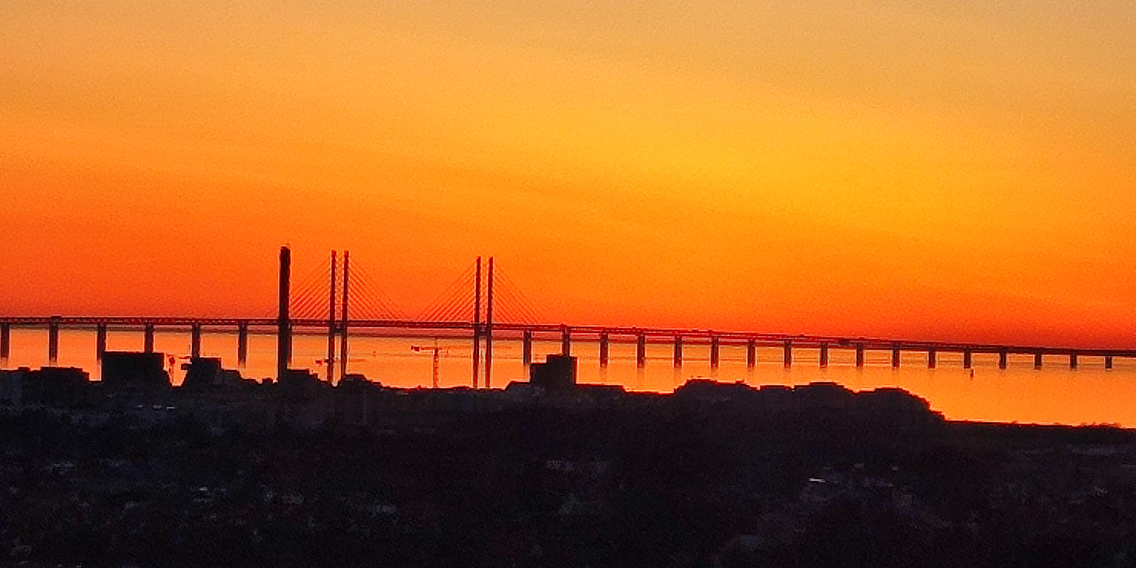 En solnedgång där himlen är alldeles orange/ röd och en bro syns i horisonten.