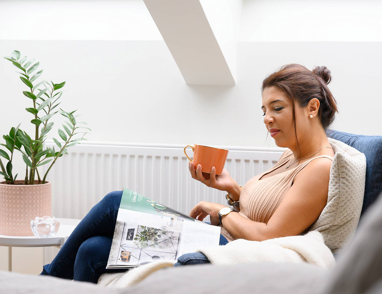En kvinna sitter i en fåtölj och dricker kaffe från en orange mugg och läser en tidning.